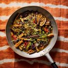 Gebratene Auberginen mit Frühlingszwiebeln und roten Chilischoten in einer Pfanne auf einem orange-weißen Stoffmuster.