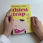 „thirst trap“ steht in großen, pinken Buchstaben auf gelbem Hintergrund. Hände halten das Buch mit einer Illustration eines Gesichts.