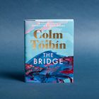 Buchcover: Der Titel "The Bridge" von Colm Tóibín. Illustration einer Landschaft mit Brücke und Bergen in Blau- und Rottönen.