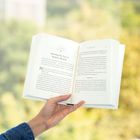 Eine Hand hält ein Buch, geöffnet bei Kapitel 51 mit dem Titel "Opening Our Eyes to Modern Miracles". Hintergrund unscharf.