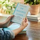 „The Prayerful Leader: A 60-Day Devotional Journey“. Hände halten das Buch, im Hintergrund Holzstapel und Pflanzen.