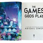 "The Games Gods Play" von Abigail Owen. Eine fantasievolle, bunte Illustration einer märchenhaften Stadt mit Wasserfällen.