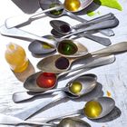 Metalllöffel mit bunten Flüssigkeiten, darunter gelb, rot und grün, angeordnet auf einer zerkratzten Oberfläche.