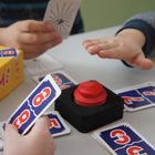 Kartenspiel mit gelber Schachtel "ALARM!", rote Taste, Kinderhände, Karten mit Zahlen und Symbolen.