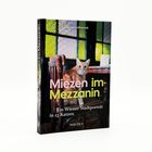 Auf einem Buchcover steht „Miezen im Mezzanin“ und „Ein Wiener Stadtporträt in 23 Katzen“. Eine orange-weiße Katze schaut aus einem Raum mit bunten Fenstern.