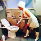 Eine Frau in weißer Kleidung, einschließlich Stiefeln und Hut, hält eine rote Bürste und ist neben einer Toilette.