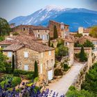 Ein Dorf aus Stein mit Zypressen auf einem Hügel, im Hintergrund ein hoher schneebedeckter Berg. Im Vordergrund Lavendelfelder.