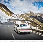 Porsche 959. Ein weißes Auto fährt auf einer kurvigen Bergstraße, umgeben von schneebedeckten Gipfeln und blauem Himmel.