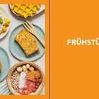 Text: "FRÜHSTÜCK". Links sind verschiedene Frühstücksgerichte auf Tellern: Waffeln, Brot und Müslischale mit Nüssen.