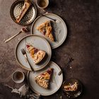 Drei Teller mit Mandel-Obstkuchen und Gabeln auf einem rustikalen Untergrund, zwei Espressotassen, ein Tuch.