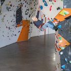 Ein Kind klettert an einer bunten Kletterwand in einer Halle. Die Wand ist mit farbigen Klettergriffen übersät.