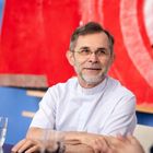 Ein Mann mit Bart in weißem Hemd lächelt. Hintergrund ein rotes abstraktes Gemälde. Glas mit Wasser auf dem Tisch.