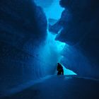 Eine Person kniet in einer blauen Eishöhle mit beeindruckenden Formationen und leuchtendem Licht.