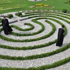 Zwei Personen in schwarzen Roben gehen durch ein grünes Labyrinth auf weißen Kieswegen.
