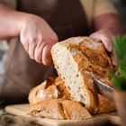 Ein Mensch mit Schürze schneidet ein frisch gebackenes Brot auf einem Holzbrett.