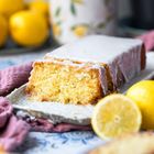 Ein angeschnittener Zitronenkuchen mit Zuckerguss auf einem Teller, umgeben von ganzen Zitronen und einem blumigen Hintergrund.