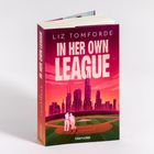 Der Roman "In Her Own League" von Liz Tomforde zeigt ein Baseballfeld vor einer Skyline bei Sonnenuntergang.