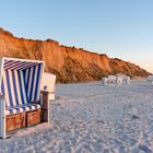 Zu sehen sind mehrere Strandkörbe auf einem sandigen Strand vor einer steilen Küste bei Sonnenuntergang.
