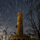 Ein hoher Ziegelturm bei Nacht, umgeben von Bäumen. Am Himmel leuchten kreisförmige Sternspuren.