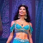 Eine lächelnde Frau in einem aufwendig verzierten, blauen Kostüm vor dekorativem Hintergrund.