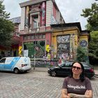Texte: "We are all", "Yes means yes", "Simplicité", "Wir machen das schon". Buntes Gebäude mit Plakaten. Frau mit Tattoos und Sonnenbrille.