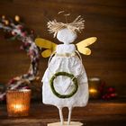 Eine Engel-Figur mit goldenen Flügeln und Heiligenschein hält einen grünen Kranz mit Herz. Im Hintergrund Kerzenlicht.