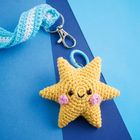 Ein gelber, lächelnder, gehäkelter Stern mit rosa Bäckchen, an einem blauen Haken und hellblau-weißem Band befestigt.