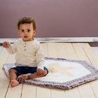 Es gibt keinen Text zu erwähnen. Ein Kind sitzt auf einer kleinen Wolldecke auf einem Holzboden, mit einem lila Hintergrund.