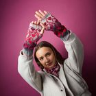 Eine Frau in grauem Mantel vor einem violetten Hintergrund trägt gestrickte fingerlose Handschuhe mit rotem Muster.