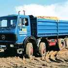 "OTTO WULF WEYHAUSEN" steht auf einem blauen Lastwagen mit Ladung. Darunter: "ERD- u. KIESWERK". Der Lastwagen fährt durch Erde.