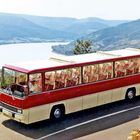 Der Text "Ikarus" auf einem roten und cremefarbenen Bus, der auf einer Straße vor einer Berg- und Flusslandschaft steht.
