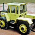 Text "MB-": Gelber Traktor mit großen, schwarzen Reifen, steht auf sandigem Untergrund, klare, kantige Kabine.