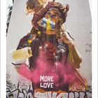 Ein Kind sitzt auf den Schultern eines Erwachsenen und malt eine Wand an. Der Text "MORE LOVE" ist sichtbar. Graffiti unten.