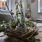 Eine Vase mit Schneeglöckchen auf einem Tisch, umgeben von einem Kranz aus Zweigen und Blättern, im Hintergrund eine Uhr.