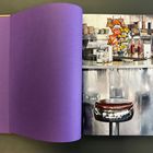 Eine lilafarbene Seite verdeckt teilweise eine Diner-Szene mit Barhockern, Snacks, Gewürzen und Ketchupflaschen auf einem Tresen.