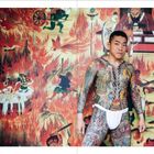 Text: "RIGHT While it seems contradictory to Buddhist beliefs..."
Ein tätowierter Mann vor einem feurigen Wandbild mit Fabelwesen.