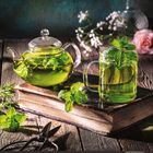 Grüner Tee in einer Kanne und einem Glas auf einem alten Buch. Minzblätter dekorieren das Getränk, umgeben von Blumen und Kräutern.