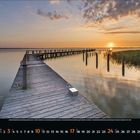 Kalender mit Tagen des Monats: 1 bis 31. Sonnenuntergang über einem See mit Steg und Wolken im Himmel.