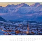 Text: "Axams bei Sonnenuntergang mit dem Wetzsteingebirge." Eine verschneite Stadt mit Kirche in einer Berglandschaft bei Sonnenuntergang.