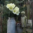 Keine Texte erkennbar. Eine Steinfigur eines Mädchens neben einem Krug mit weißen Blumen und Kiefernzweigen.