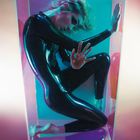 Eine Person in einem engen, schwarzen Anzug kauert in einem transparenten Kasten, umgeben von rosa und blauem Licht.