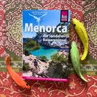 Text: "Menorca, die 'andere' Baleareninsel. Ein Reiseführer für Mallorcas kleinere, aber nicht minder attraktive Nachbarin."  
Hintergrund: Bunte Decke, drei bunte Fischfiguren.