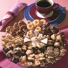 Verschiedene Kekse auf einer Platte, daneben eine Tasse Kaffee auf einem Unterteller und eine rosa Serviette.