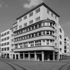 Modernes, mehrstöckiges Gebäude an einer Straßenecke. Links Fachwerkhaus. Ecke mit Straßenbahnschienen.