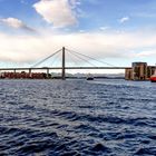 Stadtansicht mit Brücke über Wasser, rotes Schiff und bunten Gebäuden am Ufer bei bewölktem Himmel.