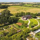 Luftaufnahme einer Landschaft mit Feldern, einem großen roten Backsteingebäude, Wegen und grünen Gärten.
