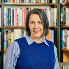 Eine Frau in einem blauen Pullover steht vor Bücherregalen voller bunter Bücher. Sie lächelt leicht.