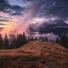 Romantische Bergszene: Eine Hütte auf einem Hügel, umgeben von Bäumen, mit einem spektakulären Sonnenuntergang im Hintergrund.