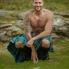 JANUAR. Ein lächelnder Mann in einem Kilt, kniend auf einer grünen Wiese, mit einem tätowierten Arm.