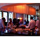 Eine Bar mit Personen an einem Tresen, gedämpftes Licht und elegante Atmosphäre. Ein Kellner serviert Getränke.